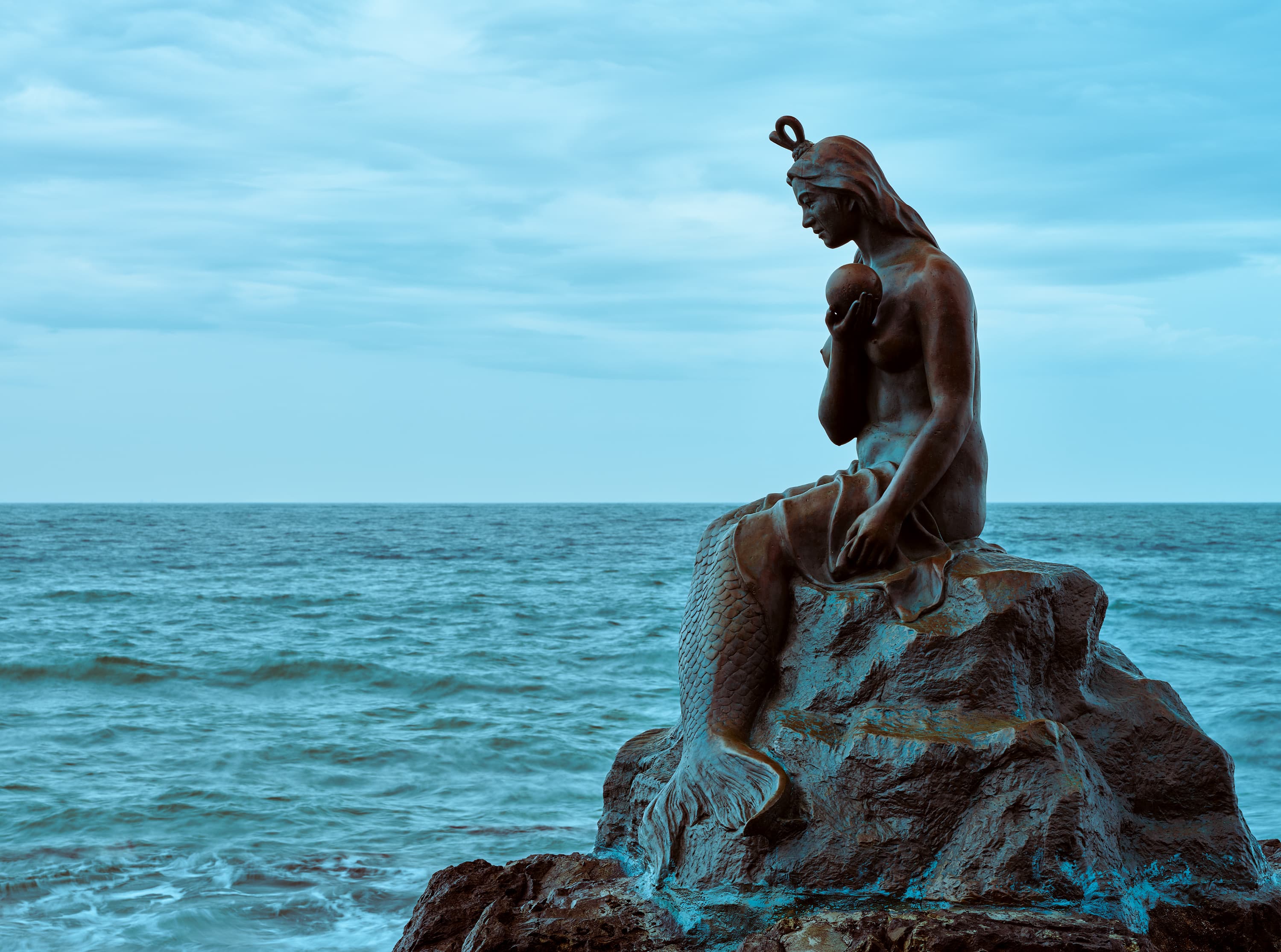 A bronze mermaid statue sitting on a rock facing the ocean under a cloudy blue sky at Dongbaekseom Island, Busan.