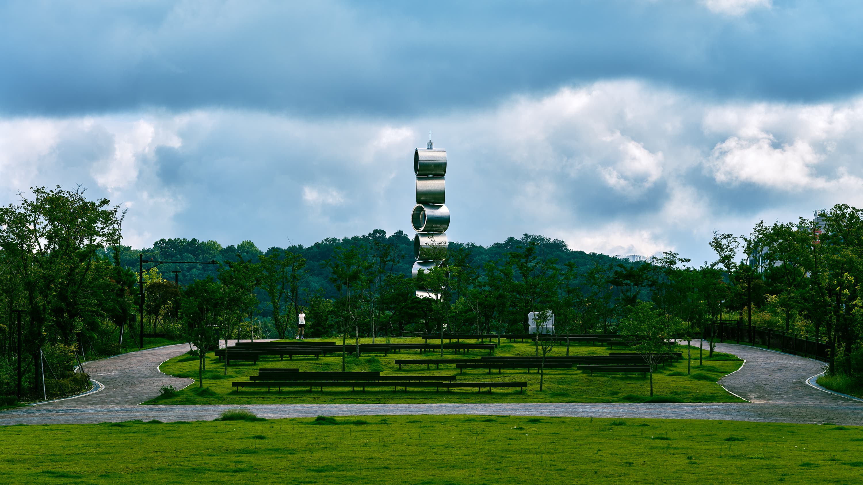 A tall modern sculpture made of stacked reflective metallic cylinders standing in the center of a green park, surrounded by trees under a cloudy sky.