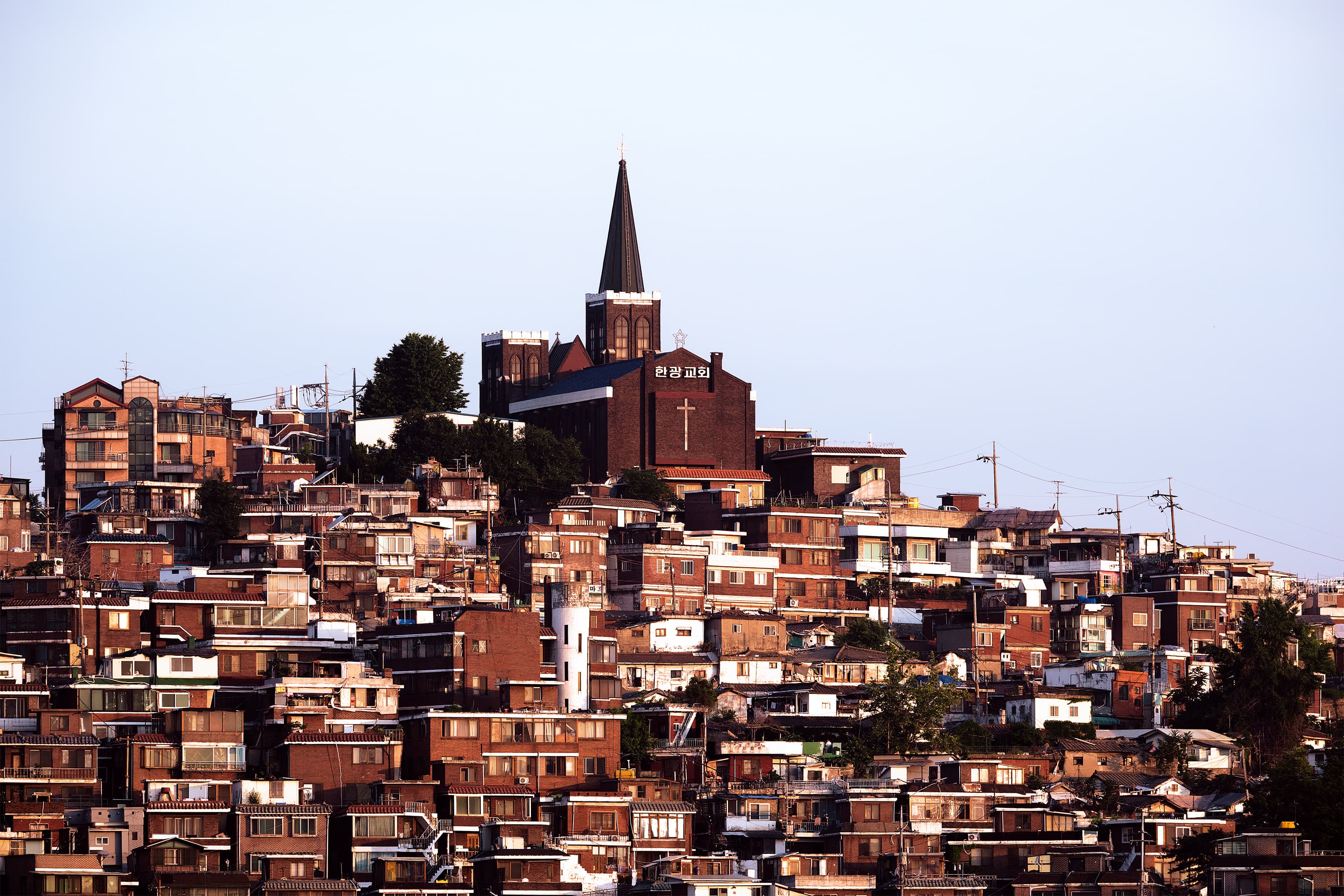 A dense hillside neighborhood of low-rise houses in warm golden light, topped by a brick church with a tall dark steeple and a cross against a pale sky.