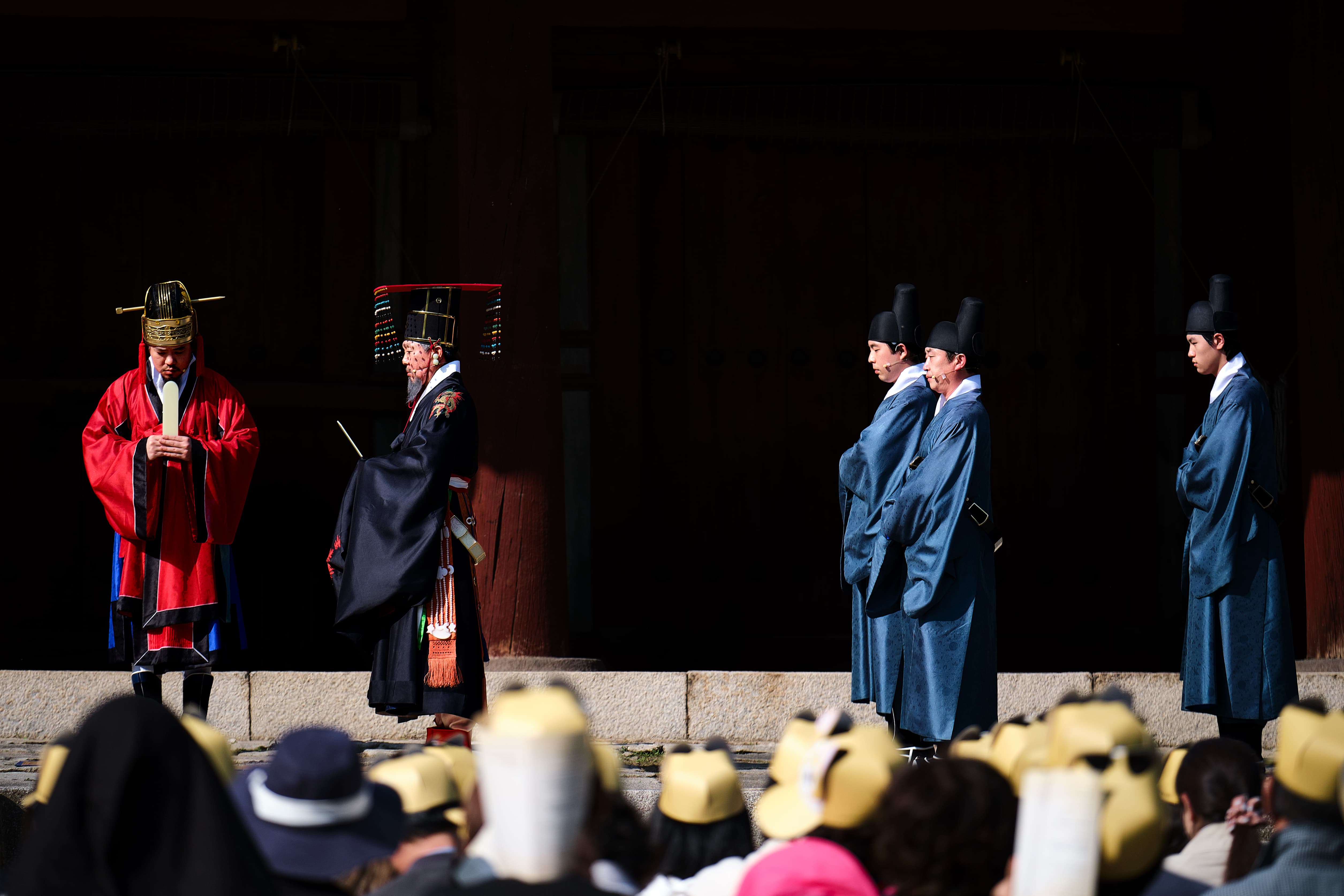 Performers in Joseon-era ceremonial robes, one in red holding a ritual tablet and others in pale blue with black hats, stand on a stone terrace before a dark wooden hall as a crowd in yellow caps watches from below.