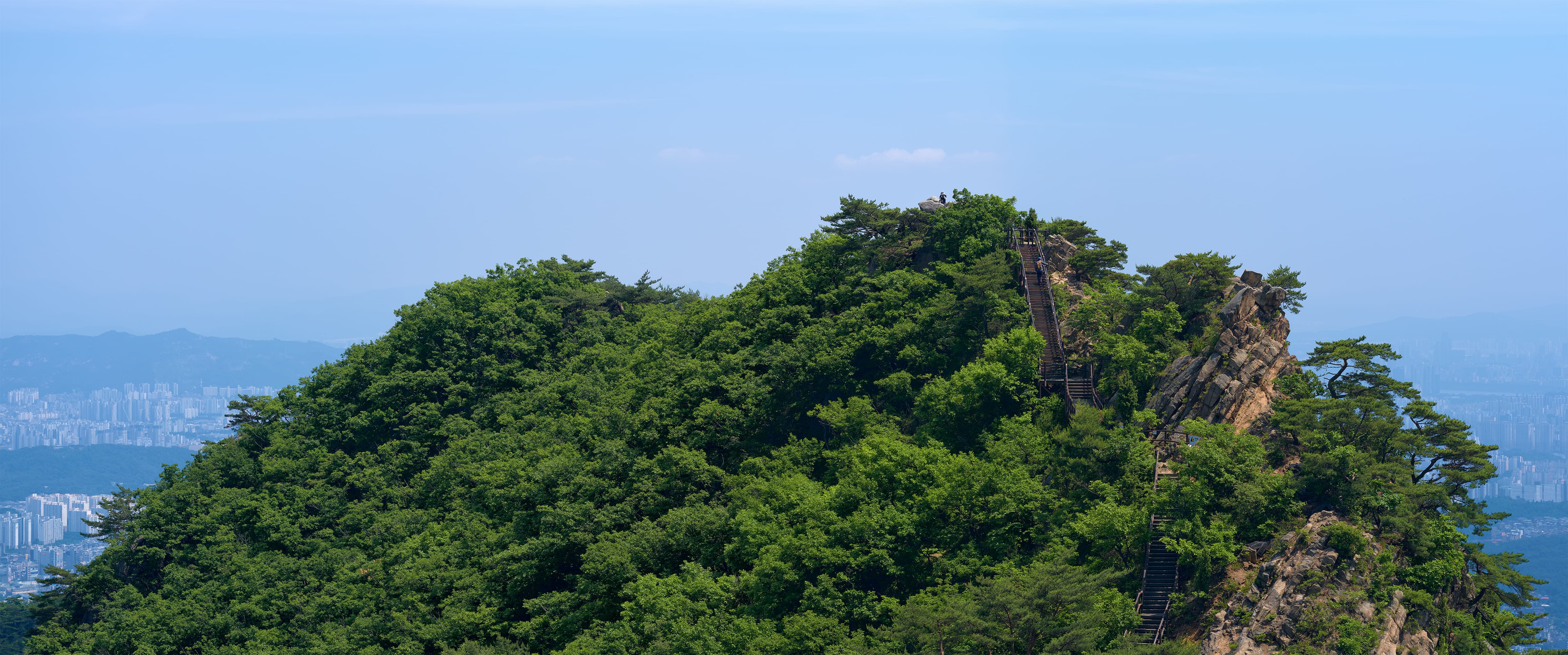 A green, forested mountain peak with granite outcrops and pines along the ridgeline, the hazy sprawl of the city visible far below under a pale blue sky.