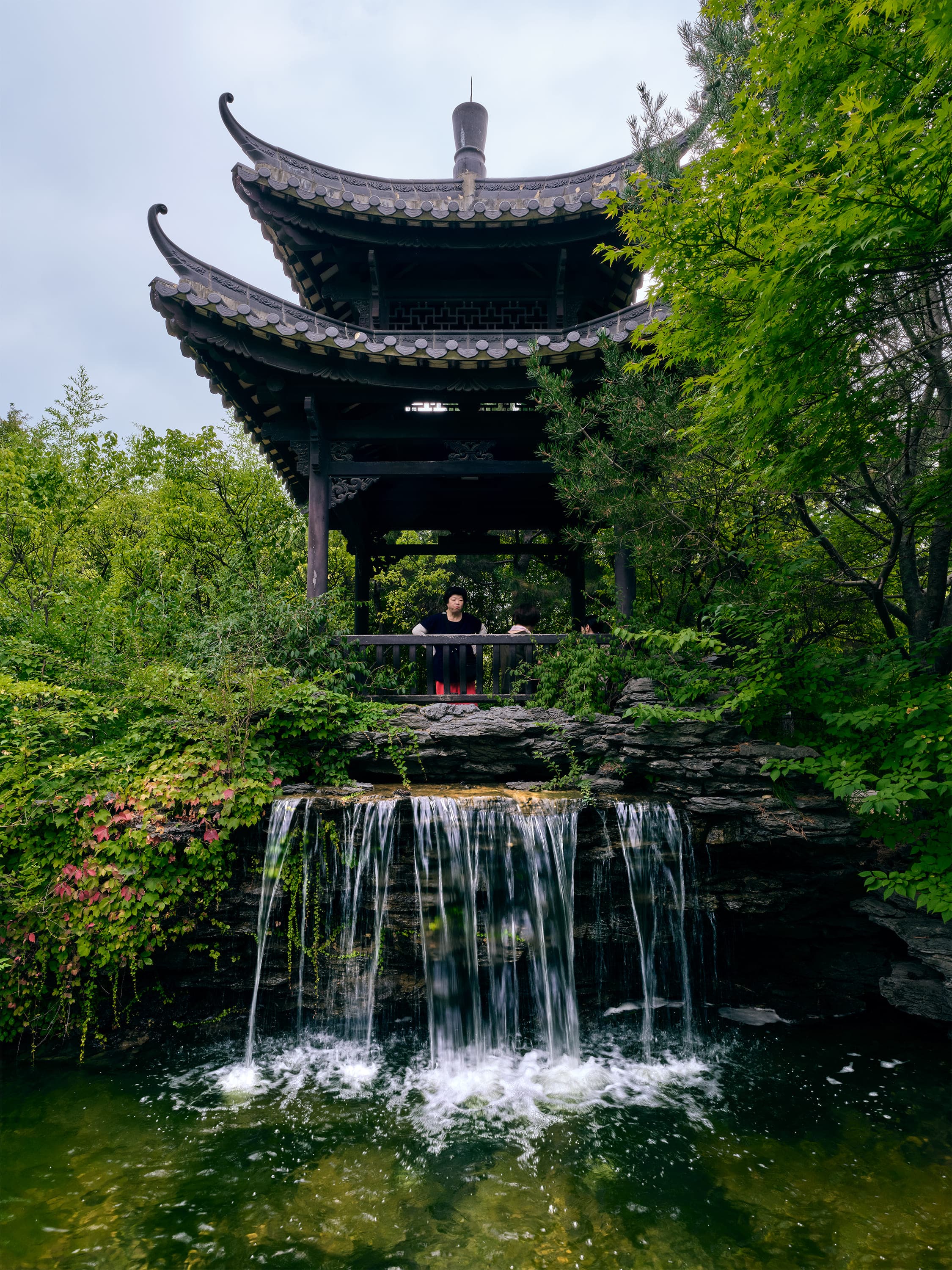 A pavilion with a steep, double-eaved tiled roof stands on a rock ledge above a waterfall that drops into a green pool, with dense summer trees on either side and a visitor in red at the railing.