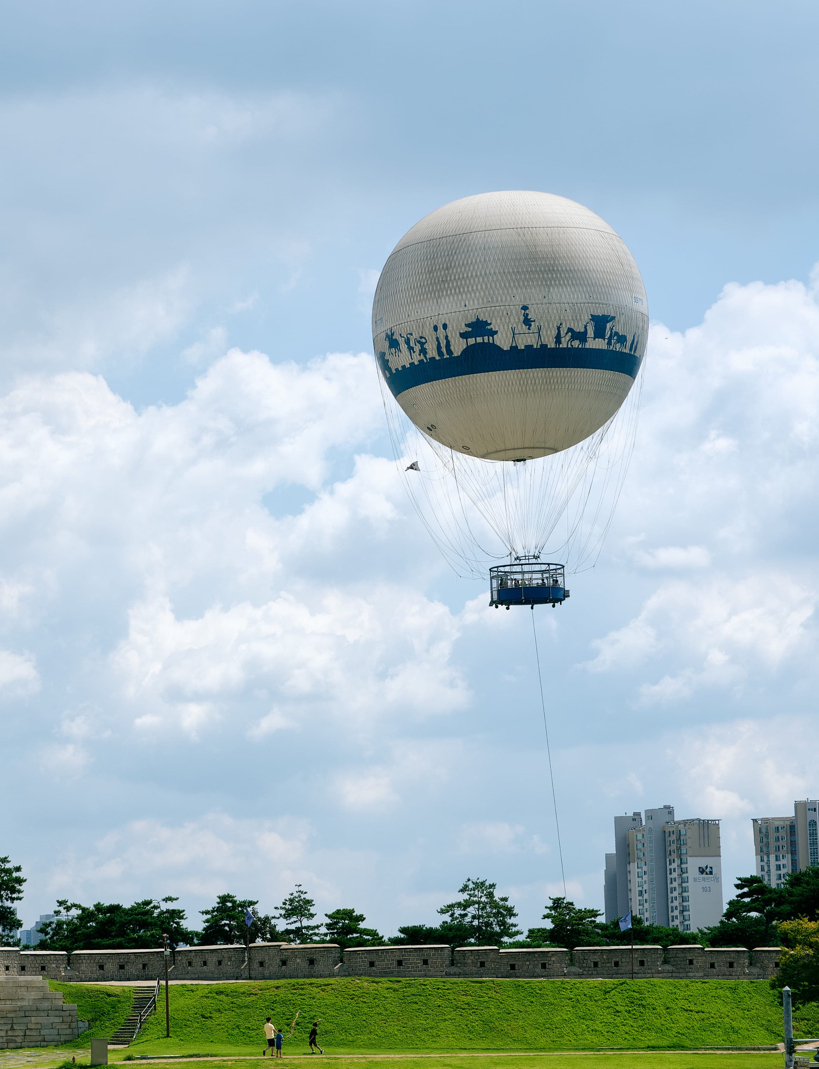 A large round sightseeing balloon floats high above a long stone fortress wall, a small basket of passengers below it, two tiny figures standing on the grass at the foot of the wall, and apartment towers in the distance.