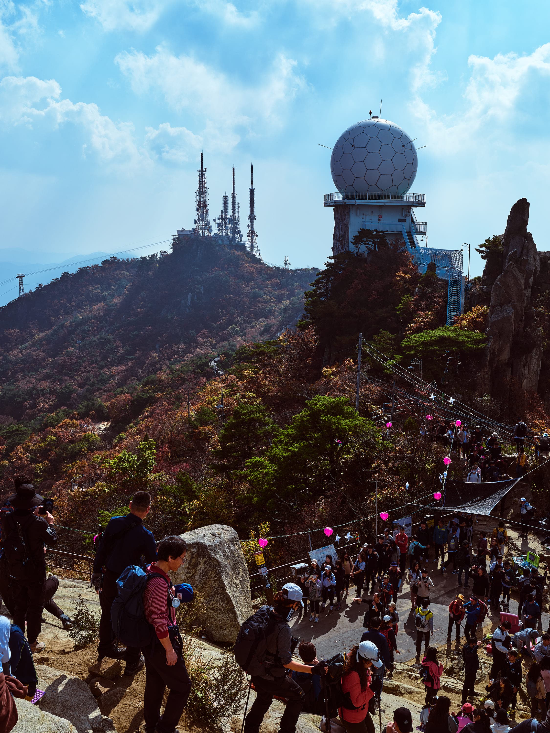 A crowded rocky mountaintop: hikers spread across bare rock and a dirt path, pink balloons and paper pinwheels strung on a line above them, a white dome shaped like a golf ball on a concrete tower, and a ridge of antenna masts behind.