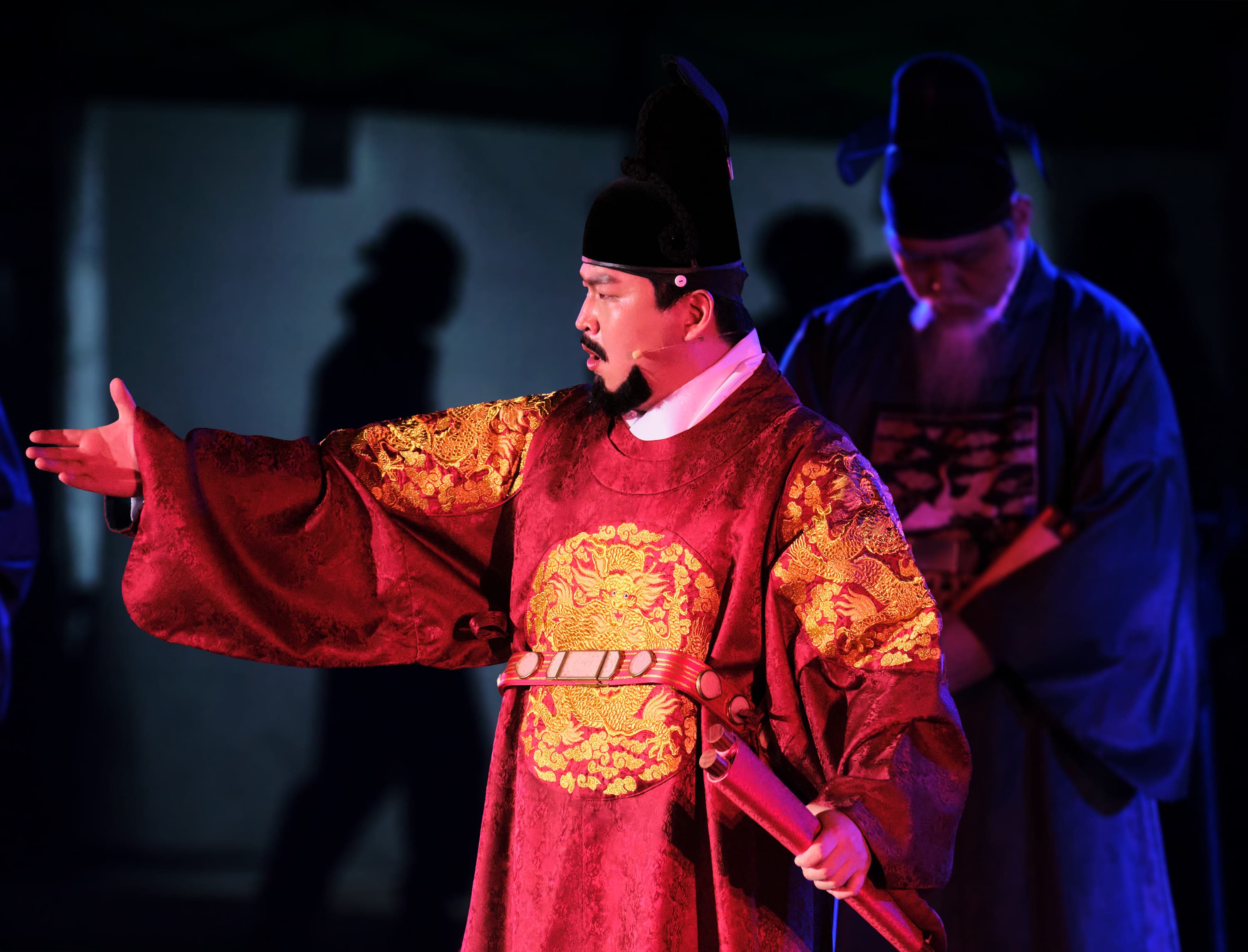 An actor on a dark stage plays a Joseon king: a red silk robe with a gold dragon roundel on the chest and on each shoulder, a black horsehair cap, a jewelled belt and a red baton in one hand, the other arm flung out as he speaks. Behind him, lit blue, an older actor in a dark official's robe stands with his head bowed.