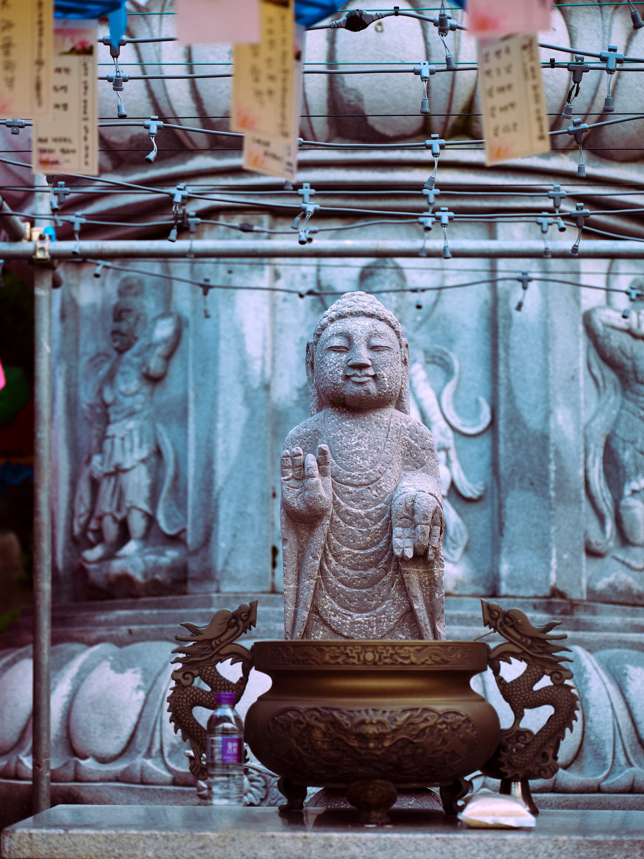 A small weathered stone Buddha stands with its right hand raised, in front of a large granite wall carved in relief with standing figures. A bronze incense burner with dragon shaped handles sits at its feet, next to a plastic water bottle. Overhead, paper slips written in Korean hang from the wires strung across the courtyard.