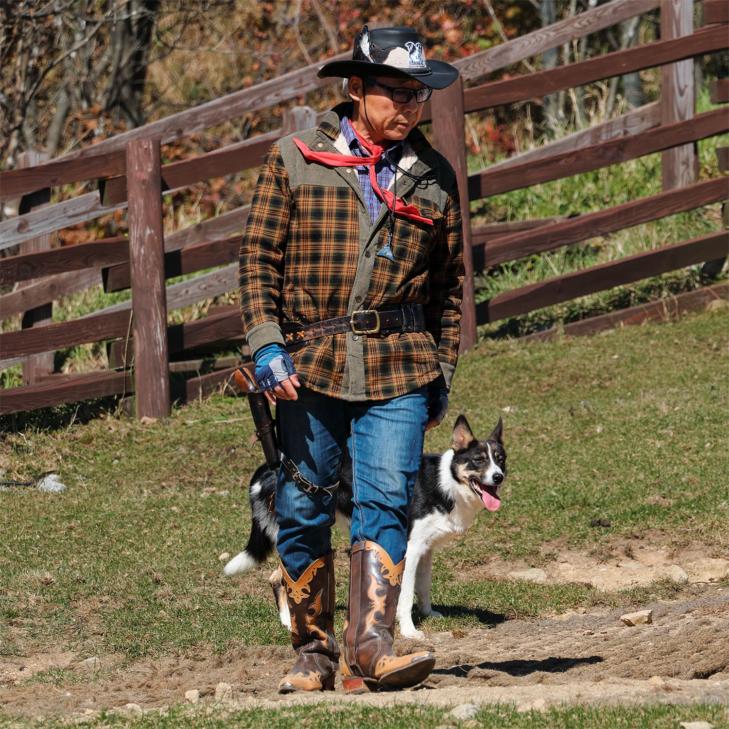 Man in cowboy attire walking with a Border Collie at Samyang Ranch, with a wooden fence and grass field in the background.