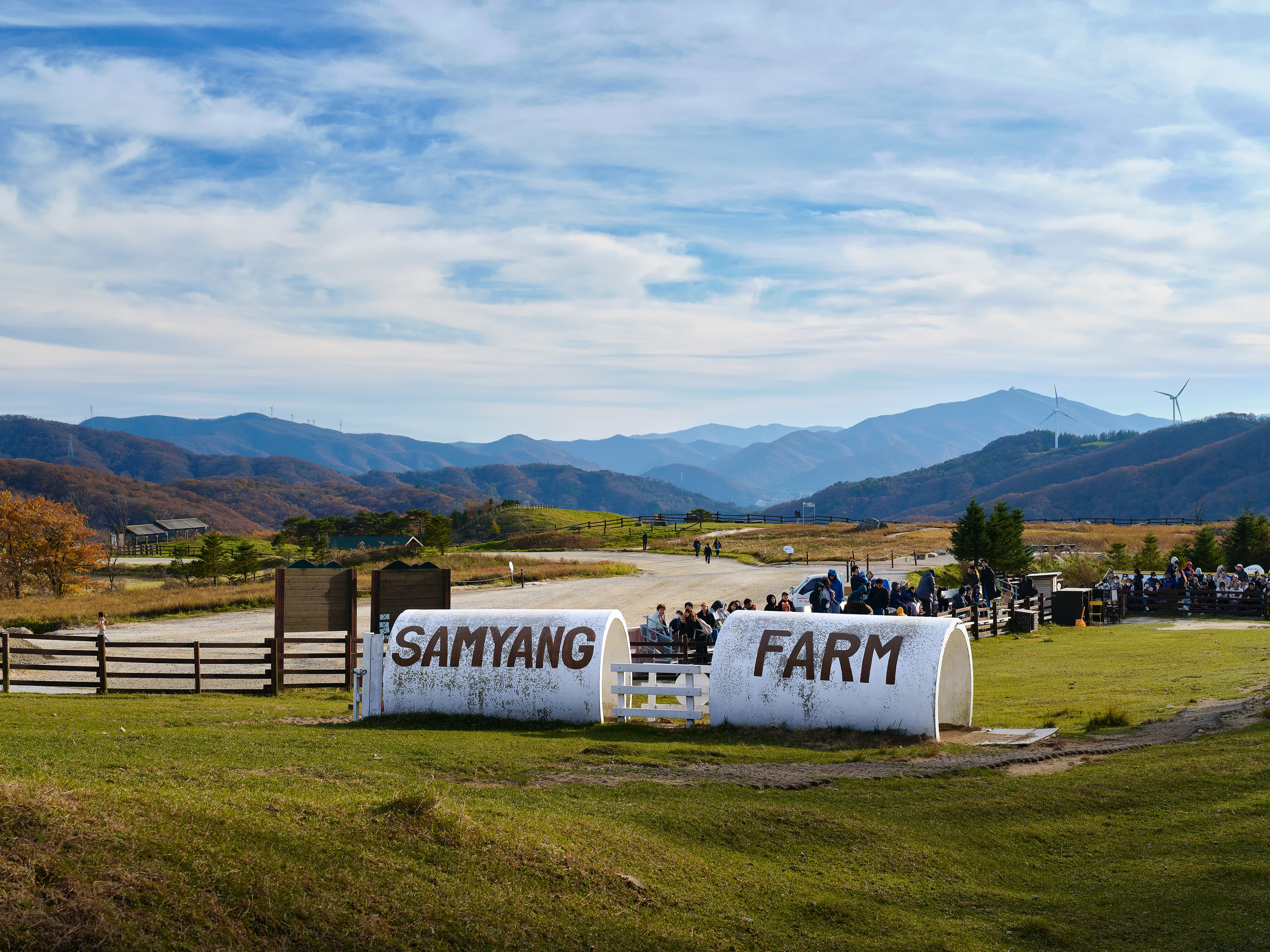 Visitors gathered at the entrance of Samyang Farm, with a white welcome sign in front and wind turbines on forested hills in the background.